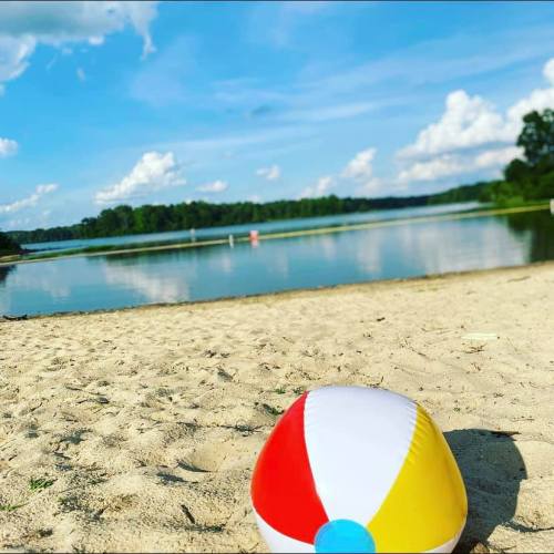 A colorful beach ball rests on sandy shore, with a calm lake and blue sky in the background.