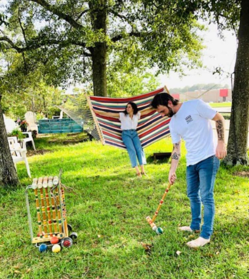 A man plays croquet on grass while a woman relaxes in a hammock under trees on a sunny day.
