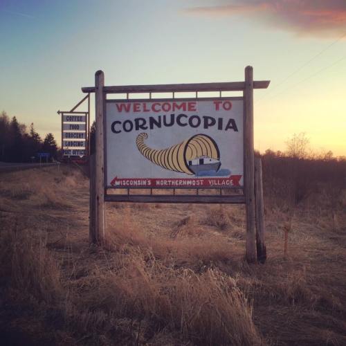 A wooden sign reading "Welcome to Cornucopia, Wisconsin's Northernmost Village," set against a sunset backdrop.