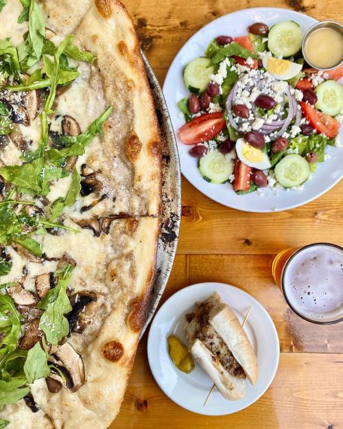 A close-up of a pizza topped with mushrooms and arugula, alongside a fresh salad and a plate with a sandwich and pickles.