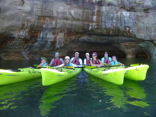 A group of people in bright green kayaks on calm water, with a rocky cave in the background.