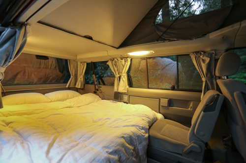 Cozy interior of a camper van with a made bed, soft lighting, and large windows surrounded by trees.