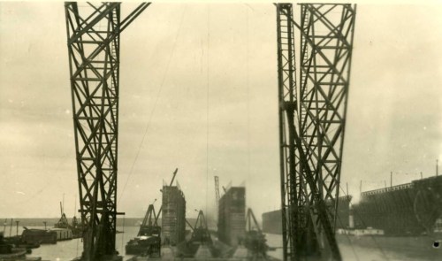 Black and white photo of a construction site with cranes and structures near a body of water.