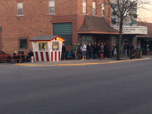 A small striped kiosk on a street corner with a line of people waiting outside a building.