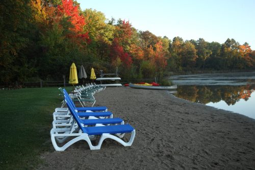 Lounge chairs line a sandy beach by a calm lake, surrounded by vibrant autumn foliage and kayaks on the water.