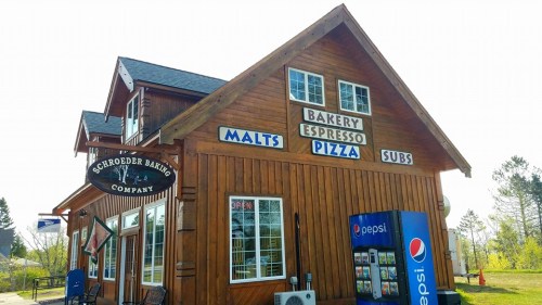 A wooden building with signs for a bakery, espresso, pizza, subs, and malts, surrounded by greenery.