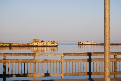 A calm waterfront scene featuring old, weathered structures on the water, with a wooden railing in the foreground.