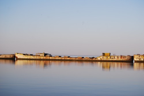 A weathered shipwreck rests on calm waters, reflecting the clear sky in the background.