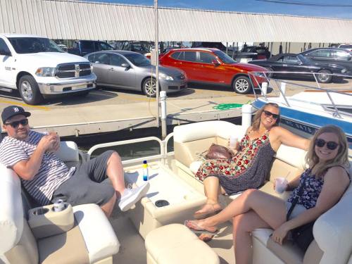 Three people relax on a boat, enjoying drinks and the sunny weather, with cars parked nearby.