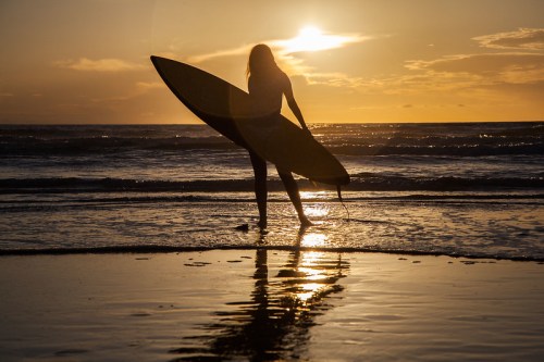Silhouette of a person holding a surfboard at sunset, standing on a beach with gentle waves.
