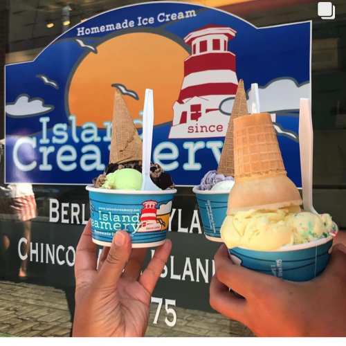 Two hands hold ice cream cones and cups in front of a colorful sign for Island Creamery, featuring various flavors.