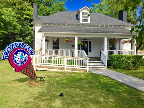A charming white house with a sign for "Frozen Cow" and colorful flower baskets on the porch, set in a green landscape.