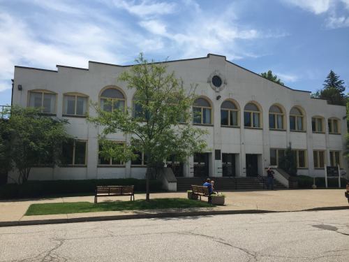 A white building with arched windows, surrounded by trees and benches, under a blue sky with scattered clouds.