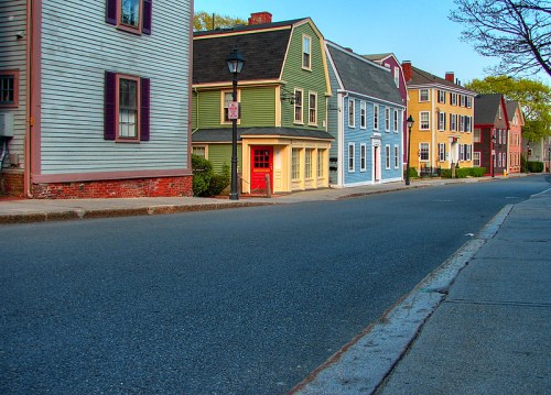 Colorful historic houses line a quiet street, with a clear blue sky above and trees in the background.