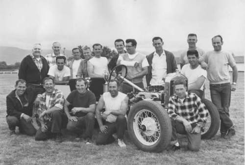 A group of 16 men poses with a vintage race car in a grassy field, smiling and celebrating their achievement.