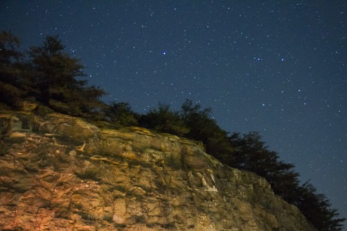 A rocky cliff illuminated by warm light under a starry night sky with scattered stars and tree silhouettes.