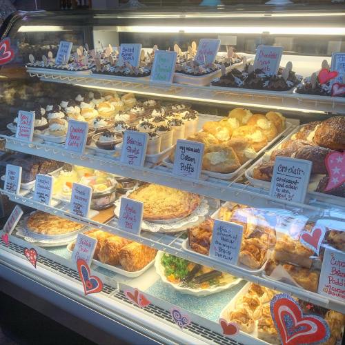 A display case filled with various pastries and desserts, including cakes, pies, and cream puffs, decorated with hearts.