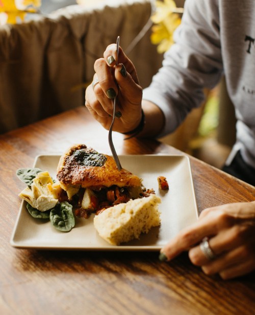 A person holds a fork, ready to eat a colorful dish with vegetables and a piece of bread on a plate.