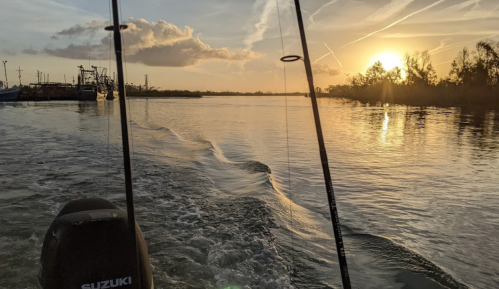 A serene sunset over a waterway, viewed from a boat with fishing rods in the foreground.