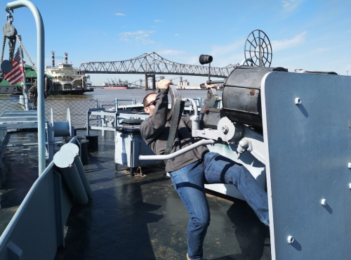 A person operates a vintage cannon on a boat, with a bridge and other vessels in the background.