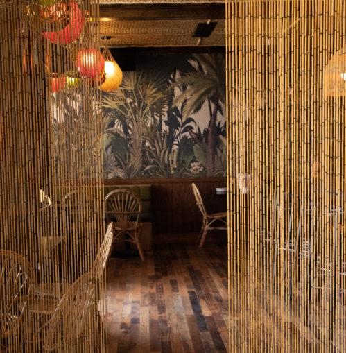 A cozy restaurant interior with bamboo curtains, wooden floors, and tropical-themed wallpaper. Warm lighting adds ambiance.