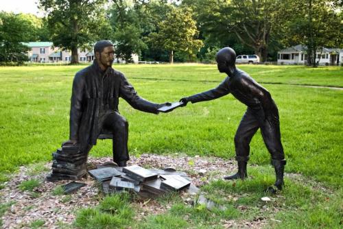 A bronze statue depicting a seated man handing a book to a standing boy, surrounded by scattered books on the ground.