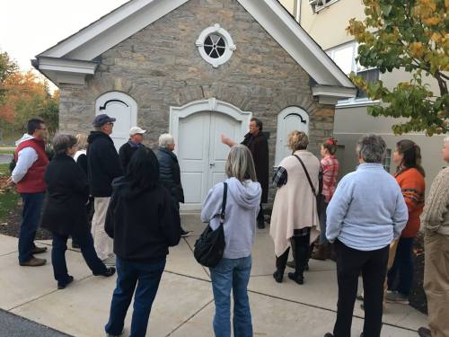 A group of people gathers outside a stone building, listening to a speaker in front of a white door.