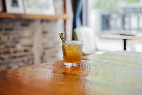 A cocktail with a sprig of rosemary sits on a wooden table in a cozy, softly lit setting.