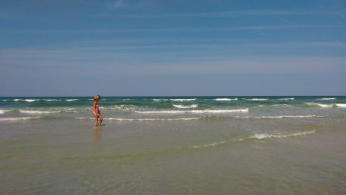 A person walks along the shoreline, with gentle waves and a clear blue sky in the background.