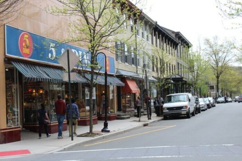 A street scene featuring shops, trees, and parked cars, with people walking along the sidewalk on a cloudy day.