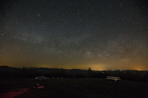 A starry night sky filled with stars and the Milky Way, overlooking distant mountains and a faint glow from a town.