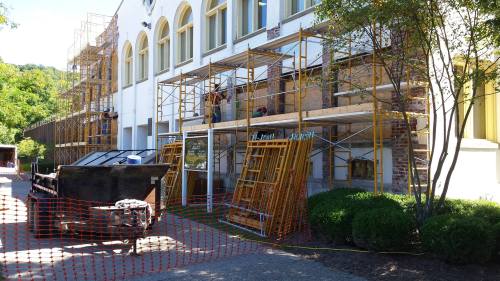 Construction site with scaffolding, workers, and equipment outside a building, surrounded by orange safety netting.