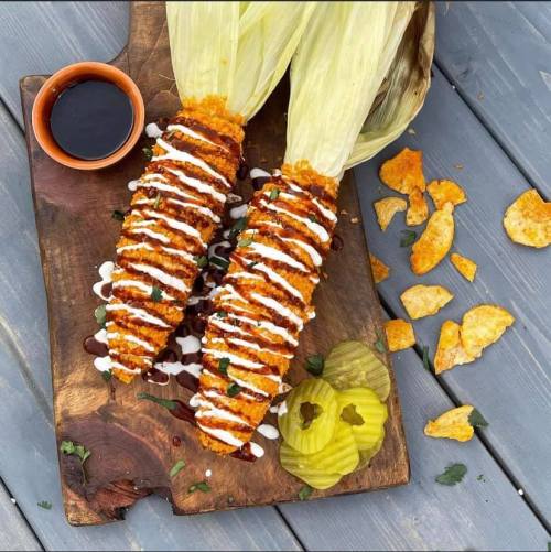 Two crispy corn on the cob topped with sauces, served with pickles and a small bowl of dipping sauce on a wooden board.