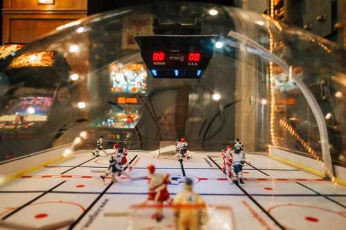 A tabletop hockey game with players on a rink, under a dome, displaying a digital scoreboard showing 0-0.