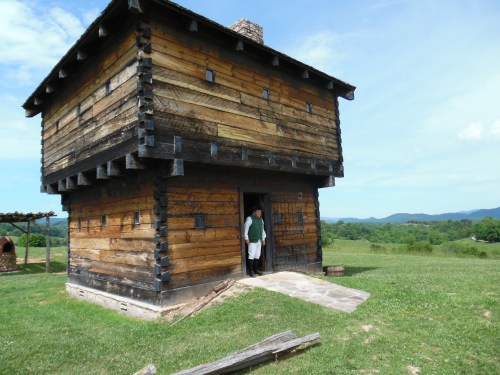 A historic wooden building with a person standing at the entrance, surrounded by green fields and mountains in the background.
