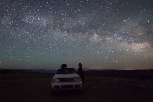 A person stands beside a parked SUV, gazing up at a starry night sky filled with the Milky Way.