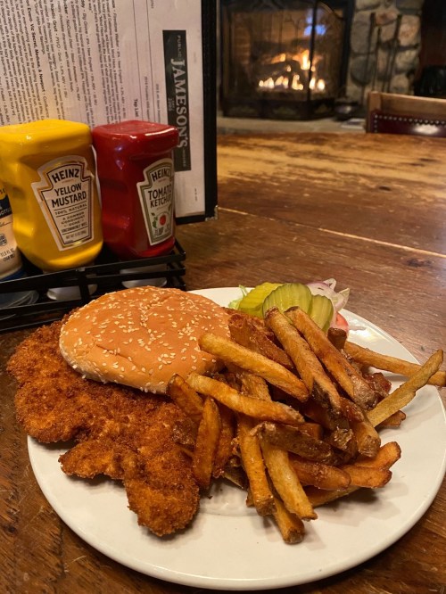 A plate of crispy fried chicken tenders, fries, a sesame seed bun, pickles, and condiments, with a cozy fireplace in the background.