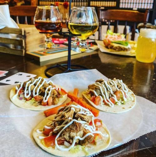 Three tacos topped with meat, tomatoes, and sauce, with drinks and food in the background on a wooden table.