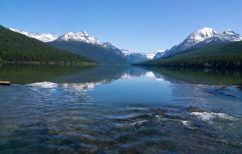 A serene lake surrounded by mountains, reflecting the clear blue sky and lush green forests. Snow-capped peaks in the distance.