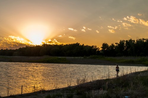 A person stands by a river at sunset, with vibrant clouds and trees silhouetted against the glowing sky.
