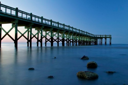 A wooden pier extends over calm waters at dusk, with smooth waves and rocks visible beneath the surface.