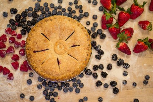 A golden-brown pie surrounded by fresh strawberries, blackberries, blueberries, and raspberries on a wooden surface.