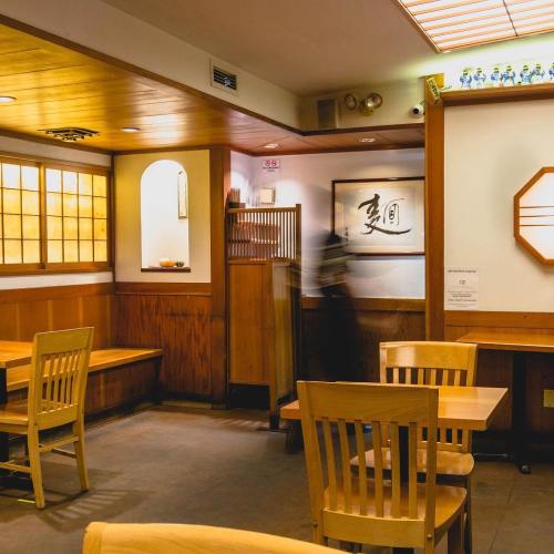 A cozy restaurant interior with wooden tables, chairs, and soft lighting, featuring a blurred figure in motion.