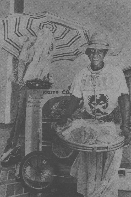 A smiling vendor stands with a colorful umbrella, selling snacks from a cart adorned with various items.
