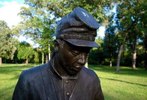 A close-up of a bronze statue of a soldier wearing a cap, set in a green park with trees in the background.