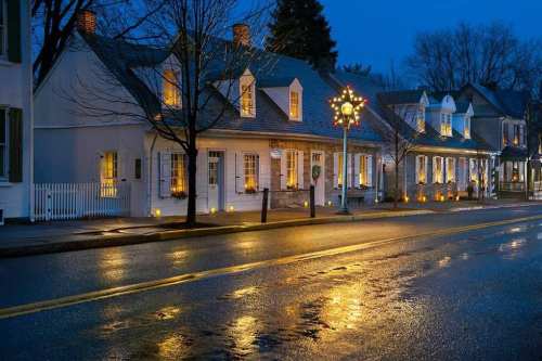 A charming street at dusk, featuring lit-up houses and a decorative lamppost reflecting on a wet road.