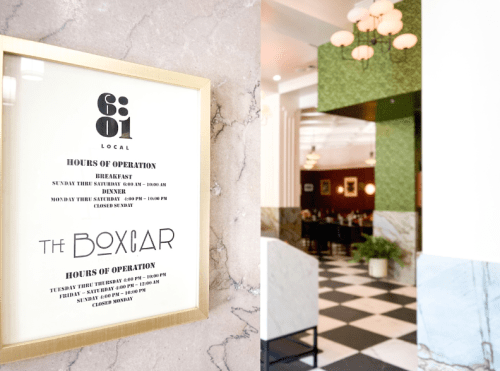 A framed sign displaying hours of operation for a restaurant, with a stylish interior visible in the background.