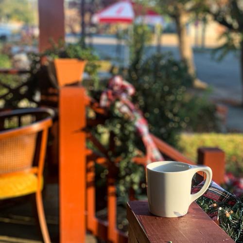 A white mug sits on a table, surrounded by greenery and outdoor seating in a cozy café setting.