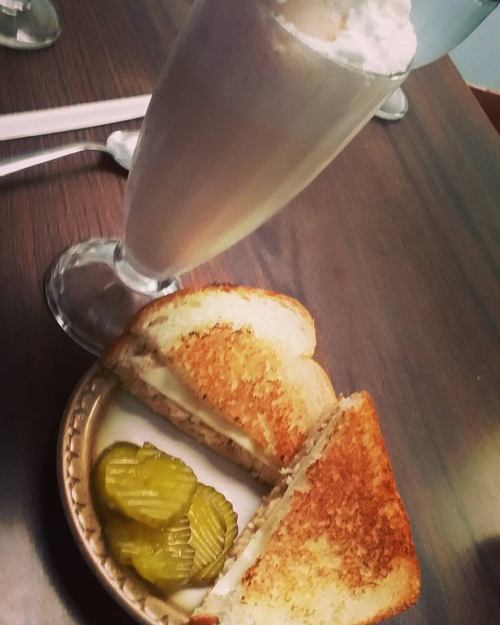 A toasted sandwich cut in half, served with pickles, alongside a tall glass of milkshake topped with whipped cream.