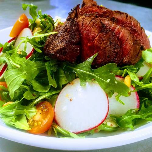 A fresh salad topped with sliced steak, mixed greens, radishes, and cherry tomatoes in a white bowl.